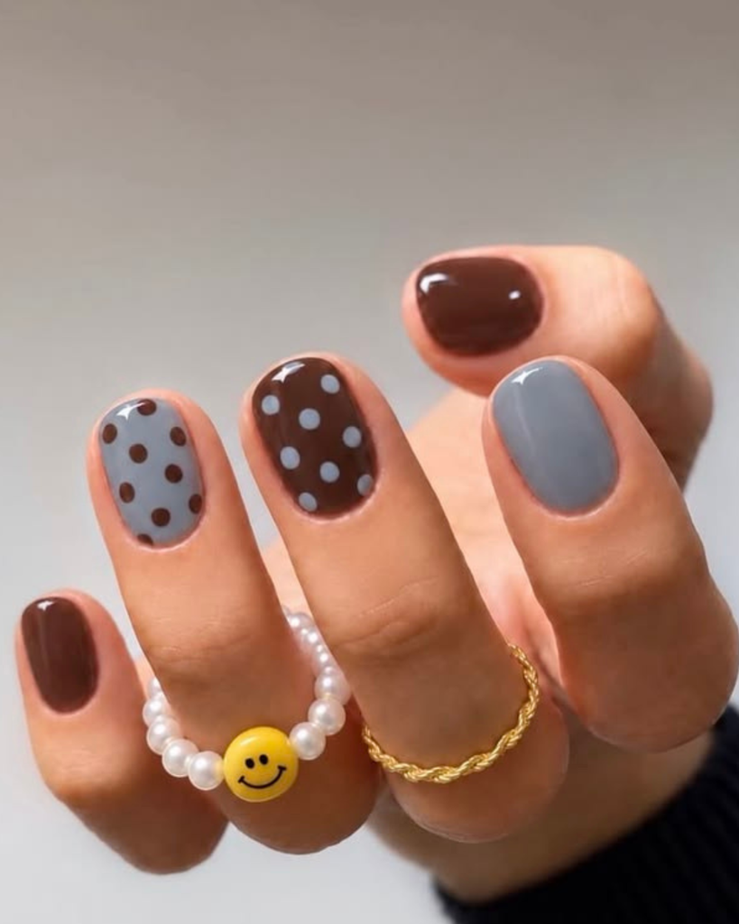 A short, rounded manicure featuring a mix of solid brown nails, a soild slate grey nail, and polka dot accent short nail design